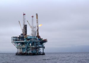 A view of an offshore oil rig in the open sea with cranes and a flare against a cloudy sky.