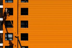 A construction worker using a cherry picker working on an orange building facade.